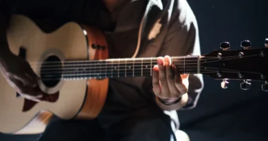 Close de uma pessoa tocando violão, focando nas mãos no braço do instrumento, iluminadas por uma luz suave ao fundo.
